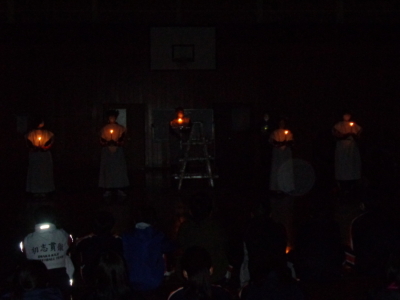 暗闇の中白い服を着た5名の生徒たちがキャンドルを持って立っている写真