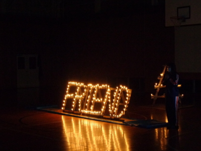 ろうそくの火が灯ったFRIENDの文字の横に女子生徒が立ってマイクを持ち話をしている写真