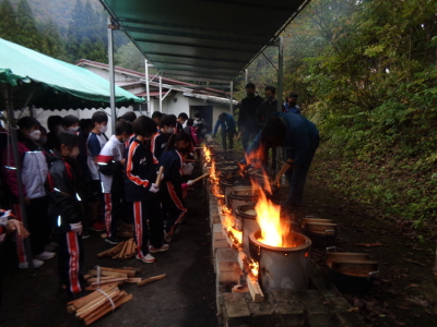 蓋をした鍋から泡が噴き出ている釜戸に生徒が並び薪をくべている写真