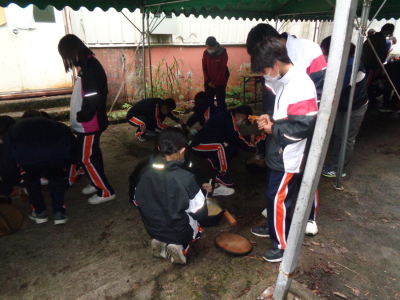 地面に豚汁の鍋が置かれ、生徒たちが周りを囲んで豚汁を注いでいる写真
