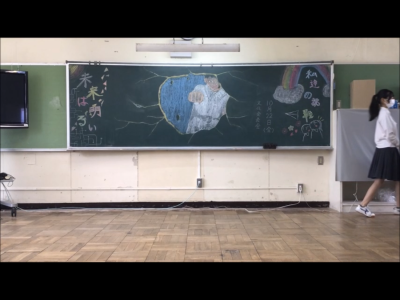 黒板にイラストと「私達の挑戦 未来は明るい」と書かれている教室の場面の映像