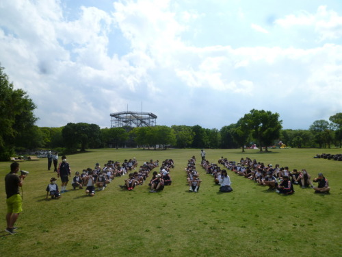 芝生の上に列になって座っている生徒と、前方で拡声器を使って話をしている男性の写真