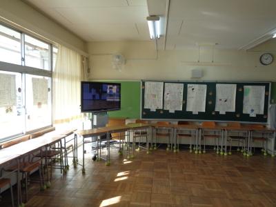 机と椅子を壁側に寄せ、教室の角に置かれたテレビに映像が映し出され、ガラス窓や黒板に模造紙が貼られている展示品の写真