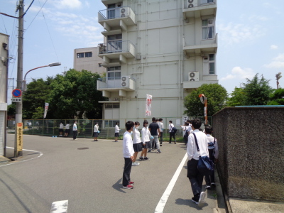 学校の近くの道路を教師や生徒が見守るなか1列になって歩いている生徒たちの写真