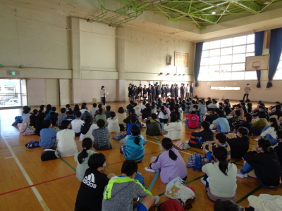 体育館の床に体育座りしている小学生の前で在校生の生徒が横一列に並んで立っている写真