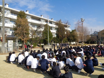 校庭に集まり座っている生徒たちが前に立っている教師の説明を聞いている写真