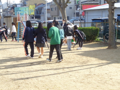 火ばさみを持った学生や緑の洋服を着た生徒など参加者たちが公園内のゴミ拾いをしている後ろ姿が写っている写真