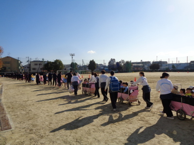 校庭を1列になってピンク色の乳母車に乗った幼児たちを運んでいる先生と生徒たちが歩いている写真