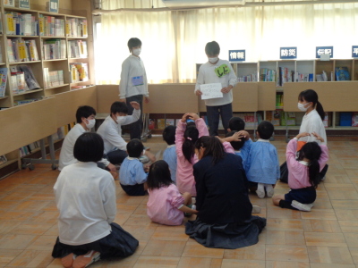 床に座っている生徒たちと園児の前で男性生徒が紙を持って話をしている写真
