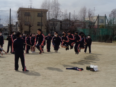 男子生徒たちが長縄を飛ぼうと高くジャンプしていて、奥では女子生徒たちが長縄跳びの様子を見ている写真