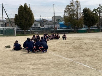 奥にサッカーゴールが見えるグラウンドの地面に生徒たちが集まってしゃがんでいる写真