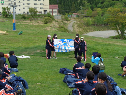 4名の女子生徒が青空の中に手を伸ばしたイラストを描いた手作りの学級旗を持って説明をしている様子の写真
