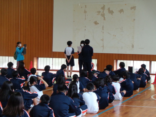 2名の男子生徒が女子生徒と向かい合って立っているのを左側の女性教師が撮影している写真