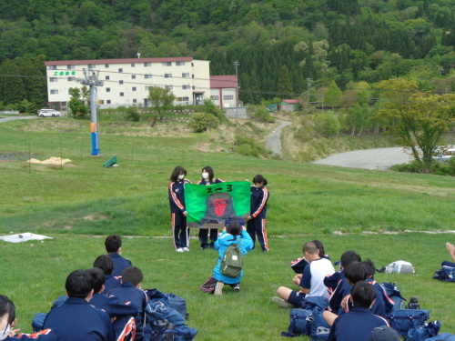 3名の女子生徒が緑色の背景に大きなサルのイラストを描いた手作りの学級旗を持って説明をしている様子の写真