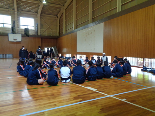 体育館の床に生徒たちが輪になって座り白色のキャップを被った男性の話を聞いている写真
