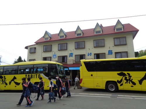 5つの三角屋根の小窓が施されている建物の前に停車したバスから降りた生徒たちが歩いている写真
