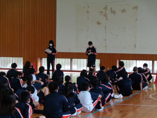 床に座っている生徒たちが前に立っている男子生徒と女子生徒の話を聞いている写真