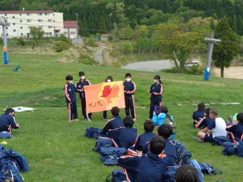4名の生徒がオレンジ色の背景に鳥の様なイラストを描いた手作りの学級旗を持って右側の女子生徒が説明をしている様子の写真