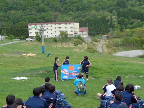 3名の女子生徒が青空の中太陽と雲のイラストを描いた手作りの学級旗を持って説明をしている様子の写真