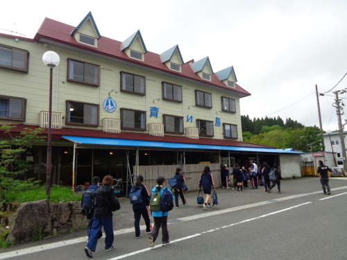 5つの三角屋根の小窓が施され壁に青い鳥と書かれた建物の下に集まっている生徒たちの写真