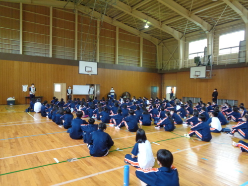 体育館の壁側に間隔をあけて立っている4名の生徒の話をクラスごとに体育座りで聞いている生徒たちの後ろ姿の写真