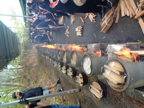 蓋をした鍋から泡が噴き出ている釜戸に生徒が薪をくべている写真