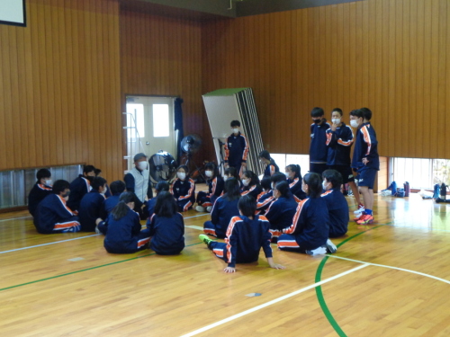 5名の男子生徒が立ち他の生徒たちが輪になって体育館の床に座りキャップを被った男性の話を聞いている写真