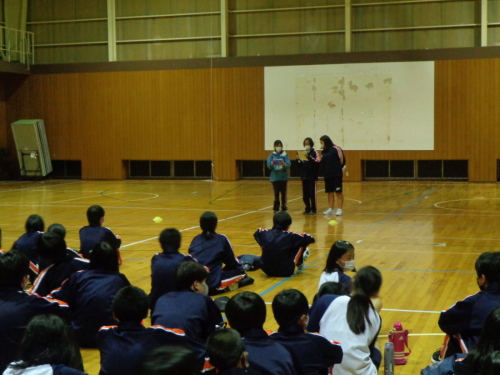 前に立っている3名の女子生徒の話を床に座って聞いている生徒たちの後ろ姿の写真