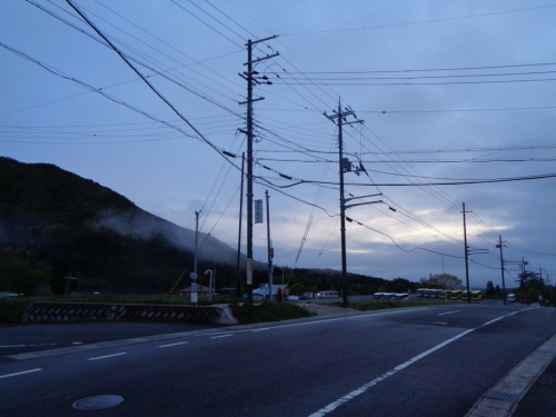 曇りがかっている小高い山の奥から太陽の光が薄っすら差し込んできそうな宿泊施設前の坂道付近を撮影した写真