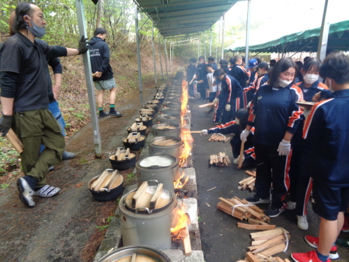 蓋をした鍋から泡が噴き出ている釜戸に生徒が薪をくべている写真