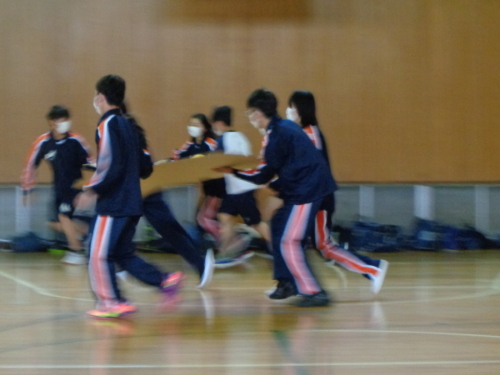 4名の生徒が大きなダンボールの上に黄色のボールを乗せて走って競っている写真