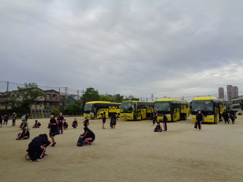 グラウンドの奥に黄色の大型バスが4台停まっており、そのバスの前で生徒たちが荷物を持ち、少しずつ集合している様子の写真