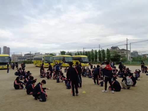 黄色の大型バスの前で、生徒たちが荷物を持って整列しようとしている写真