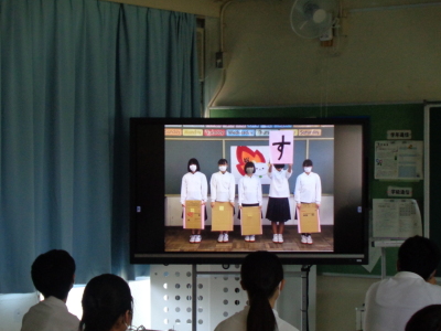 5名の女子生徒が横一列に並び右から2番目の女子生徒がダンボールに貼られた大きな「す」の文字を見せているのがうつっている、テレビ画面を生徒たちが見ている写真