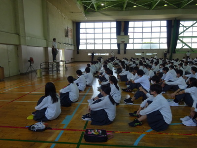 体育館の床に体育座りしている生徒たちが檀上に上がっている男性教師の話を聞いている写真