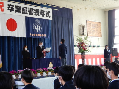 卒業証書授与式と書かれた幕がかかっている体育館のステージに立つ男子生徒の前で卒業証書を読み上げている校長先生の写真
