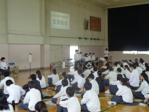 スクリーンが映し出された壁の下に設置されたマイクの前に立つ男子生徒の話を体育座りをして聞いている生徒たちの後ろ姿の写真