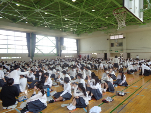 体育館内で体育座りしている生徒たちが手を挙げている人数を数えている写真