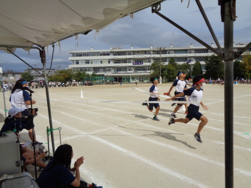 ゴールテープを切りながら走る赤組の生徒、すぐ後ろを走る緑、青、オレンジの組の生徒達が写っている写真