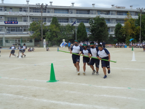 緑の組の女子生徒2人と男子生徒2人が緑の長い棒を持ち、コーンへ向かって走っている写真