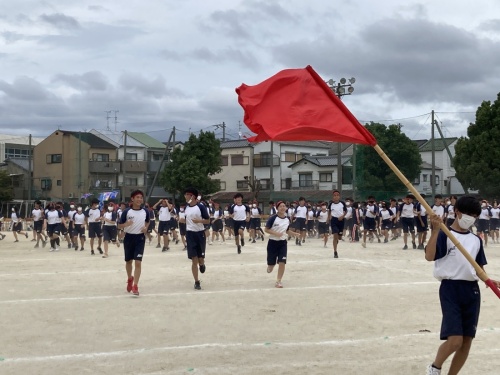 赤組の応援旗を持った生徒の後ろを赤組の生徒たち全員が走っている写真