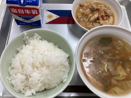 フィリピンの小さな国旗が置かれたトレイに並べられたごはん・牛乳・焼豚スープ・アドボの給食の写真