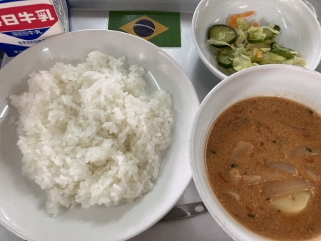 小さなブラジルの国旗が置かれたトレイに並べられたごはん・牛乳・ムケッカ・フレンチサラダの給食の写真