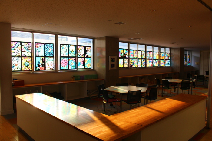 太陽の光がさす教室の窓に、ステンドグラスの作品が飾られ、机や棚の上が淡く色づいている写真