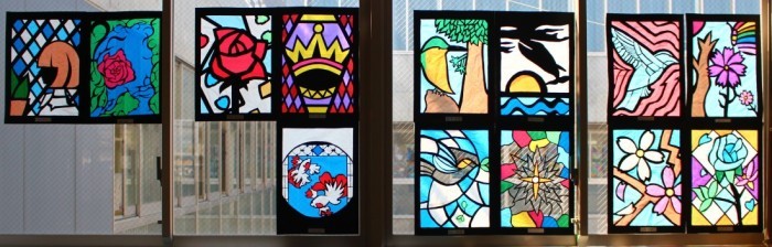 鳥や花、女性の横顔、幾何学模様が描かれた13枚のステンドグラスの作品が窓に飾られている写真