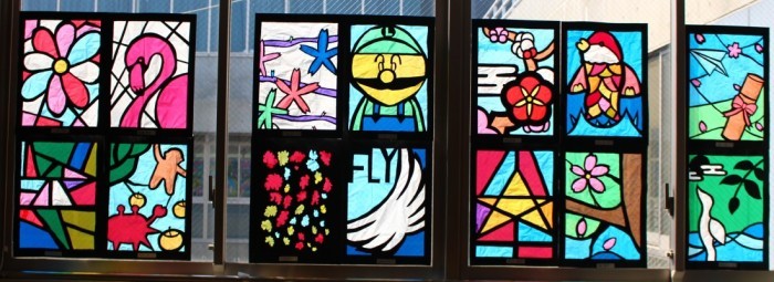 太陽が当たる窓に、花、鳥、キャラクターなどが描かれ14枚のステンドグラスの作品が飾られいる写真