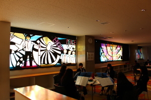 鳥や男の子が描かれた大きなステンドグラスの作品が飾られた室内で、着席している学生が手元の書類を見ている写真