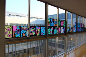 廊下の窓にカラフルな木や花などが描かれたステンドグラスの作品が飾られている写真
