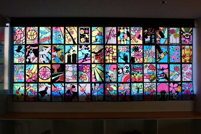 制服姿の学生や花や鳥などが描かれた56枚のステンドグラスの作品が飾れている写真