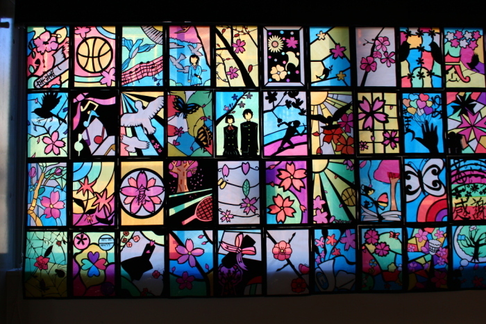 学生服をきた男女、桜の木や幾何学模様などが描かれた40枚のステンドグラスの作品の写真